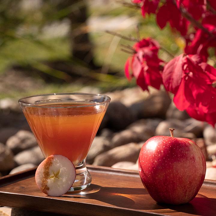 Caramel Apple Martini next to a red apple on a wood board near fall foliage 